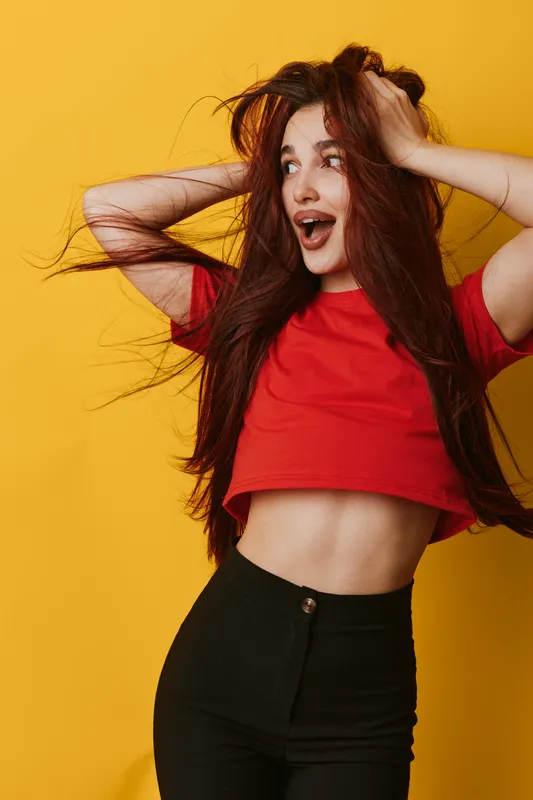 Young woman with long hair and red shirt posing excitedly against yellow background. Expressive face, casual outfit with black pants, lifestyle and happiness in studio portrait.