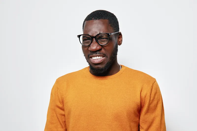 African American man with dark skin wearing glasses and orange sweater grimacing with eyes closed on white background. Portrait, emotion, facial expression and casual clothing indoors.