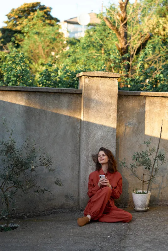 A woman in a red dress sits on the ground in a sunlit garden courtyard, looking at a smartphone with a calm, contemplative expression, representing relaxed female lifestyle