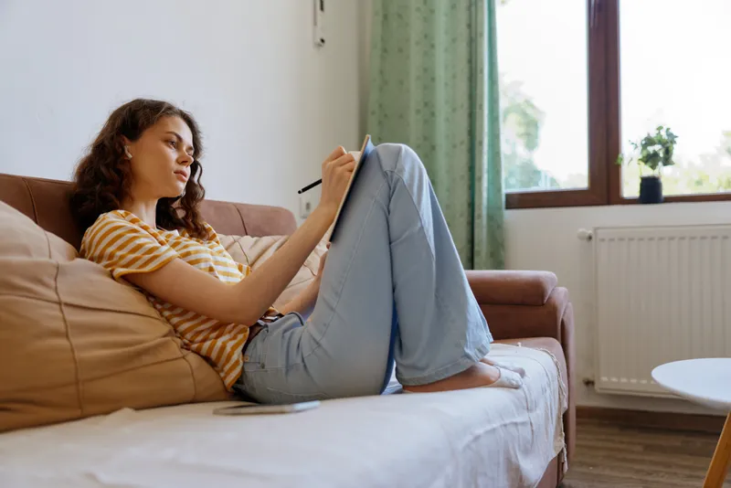 Young woman sits comfortably on a soft sofa at home, writing on a tablet with relaxed posture, smiling softly as she focuses on her ideas in natural light from a nearby window, wearing a casual