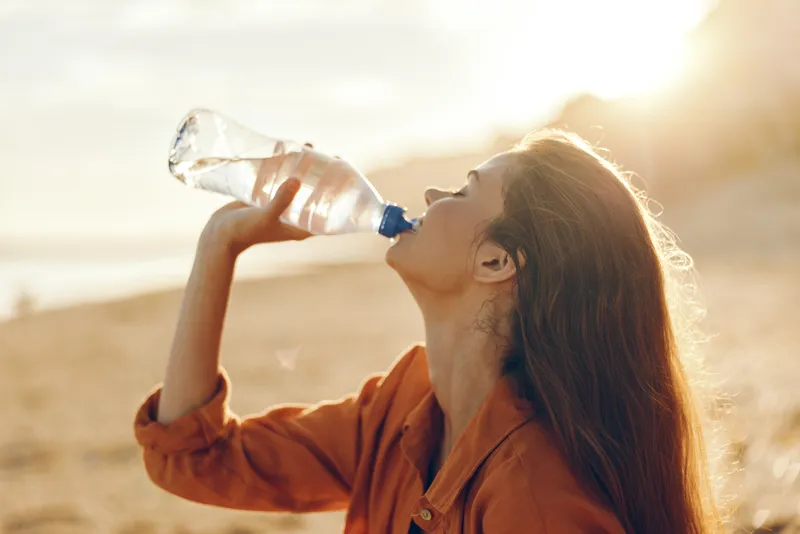 Woman drinking water from bottle outdoors, summer beach background, sunlight flare, relaxed posture in profile, warm wind, healthy hydration moment, natural colors, soft breeze and bright sky