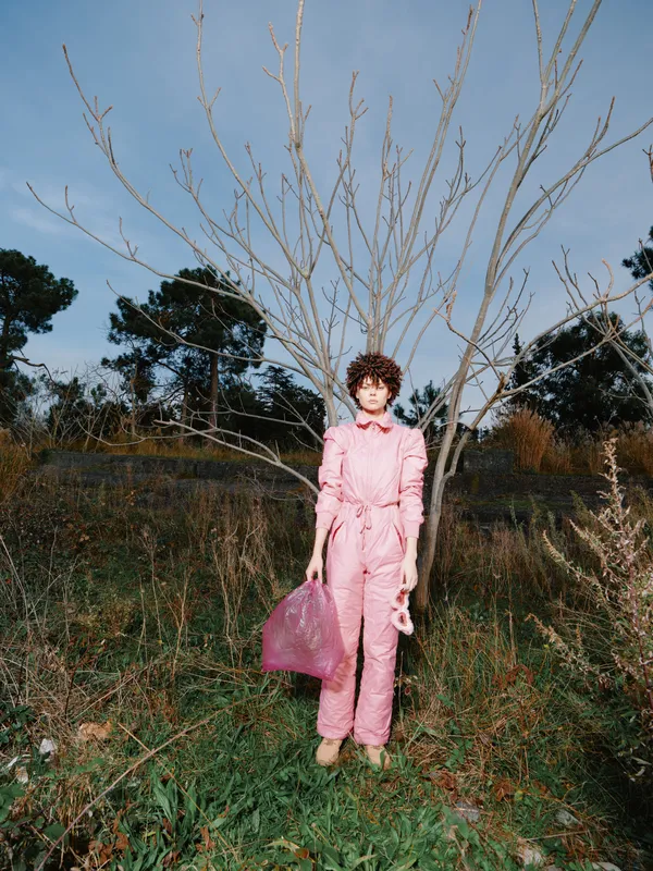 Woman in pink jumpsuit standing in a natural field with bare trees, tote bag, and clear sky, showcasing fashion in an outdoor setting with calm mood and simple silhouette