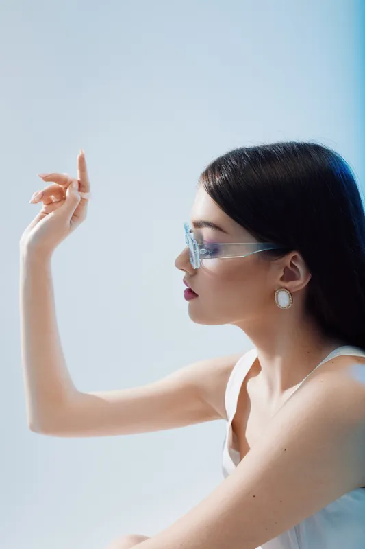 Elegant woman in transparent glasses poses with raised arm against a light background. Her smooth skin and stylish earrings highlight beauty and fashion concepts.