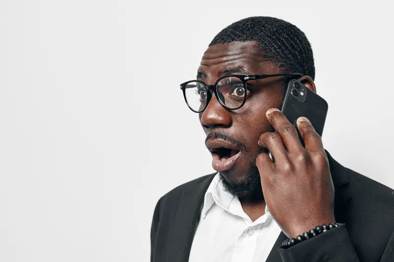African American man with dark skin wearing glasses and black suit talking on smartphone with surprised expression isolated on white background in studio portrait.