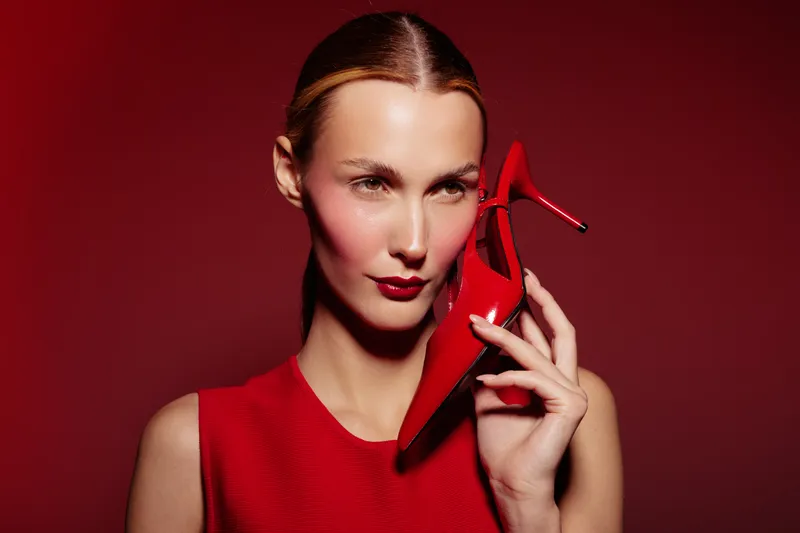 Stylish woman in red dress holds red high heel shoe near face against red background. Fashion concept with bold makeup and elegant pose in studio lighting for glamour and style.