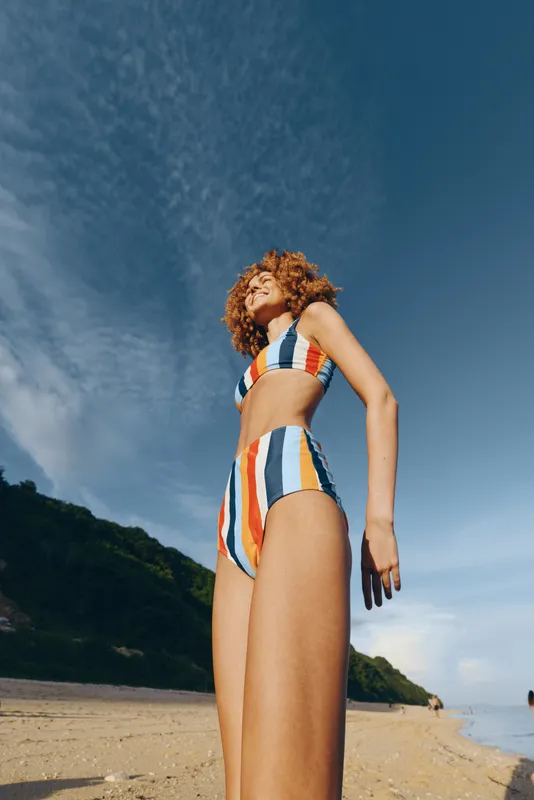 Woman standing on beach in striped swimsuit during summer, sunlight and clear sky behind her, outdoors vacation mood, relaxed posture on sandy shoreline, nature and ocean scenery, vertical portrait