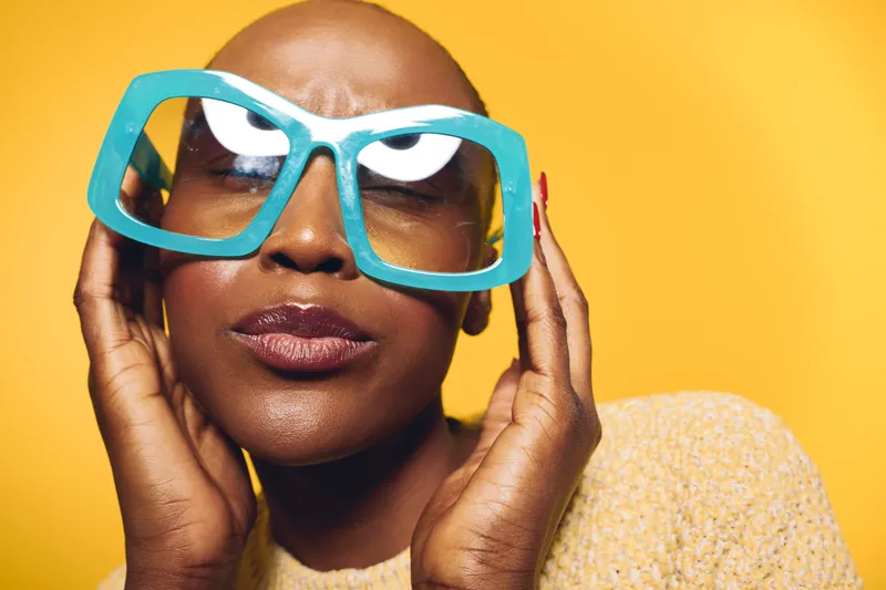 Portrait of a confident black woman with dark skin wearing oversized blue glasses and a yellow sweater against a bright yellow background. Concept for fashion and style.