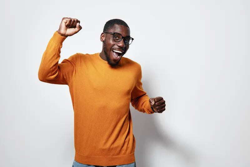 African American man with dark skin wearing orange sweater and glasses celebrating with raised fist and smiling on white background. Happy, excited, casual style.