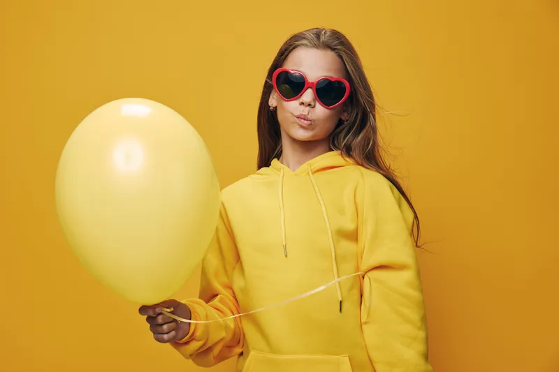 Young woman with long hair wearing yellow hoodie and heart shaped sunglasses holding yellow balloon on bright yellow background, casual and fun portrait.