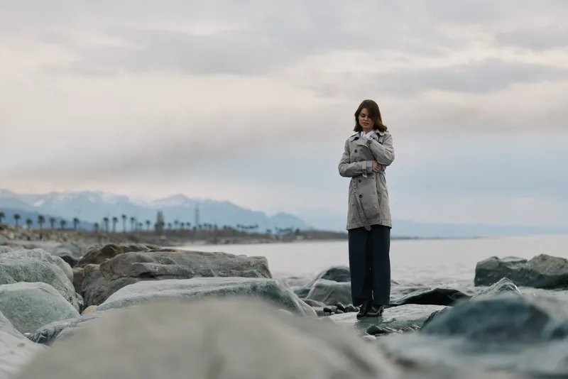 A woman in a patterned coat stands on rocky shore near calm sea under cloudy sky. Concept of solitude, nature, and cold weather outdoors with distant mountains and palms in background.