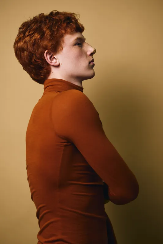 Profile of young man with red hair, wearing a rust-colored turtleneck sweater, standing against a plain beige background with folded arms, serious expression, and side view portrait.