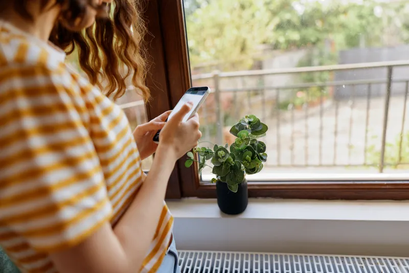 Woman sits by a sunny window, holding a smartphone in a casual home setting, striped shirt, plant on the sill, soft natural light, calm everyday moment for lifestyle photography