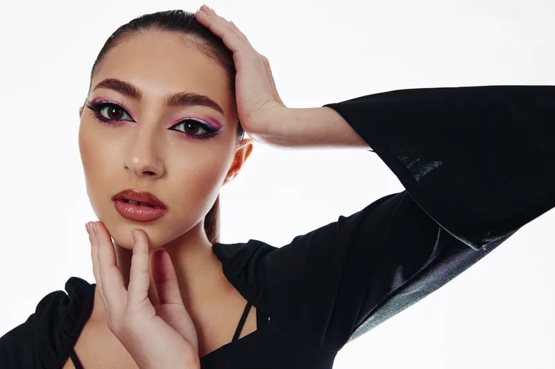 Close-up of a beauty creative fashion model with bold pink eye makeup and glossy lips wearing black clothing. The model poses against a white background, creating a stylish, modern look.