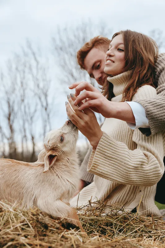Lifestyle couple outdoors with a gentle goat kid, feeding and petting, warm sweaters, soft autumn light, candid rural moment showcasing affection and connection with animals