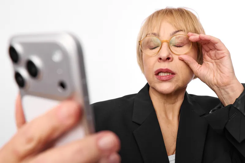 Stressed woman in business attire holds glasses and rubs tired eyes while checking a smartphone. Blurred phone hand suggests anxiety and pressure under stress, work fatigue.