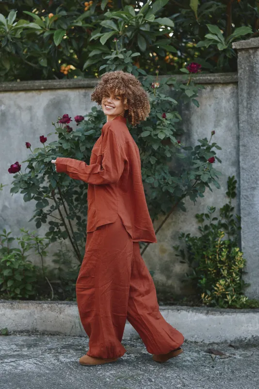 Young woman with curly hair walks along a garden path in an orange loose jumpsuit, smiling softly beside a weathered wall and blooming plants during a sunny afternoon