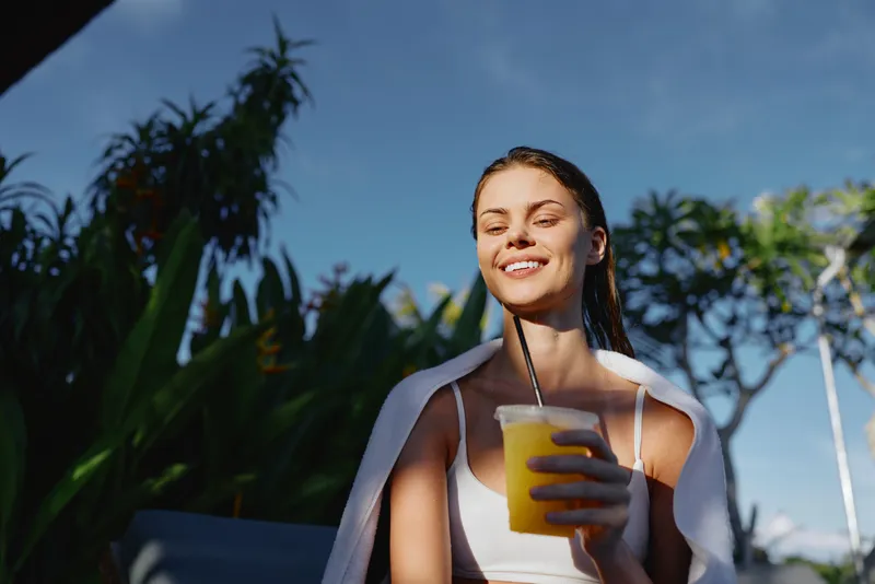 Smiling woman outdoors enjoying fresh juice, relaxed posture, sunlit skin, casual summer outfit, tropical plants background, natural lifestyle moment.