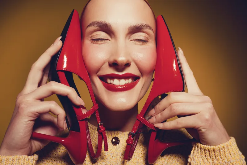 Happy woman with closed eyes holds red high heel shoes near face smiling. Concept of fashion, style, beauty, and joyful emotions in studio with warm lighting and cozy sweater.