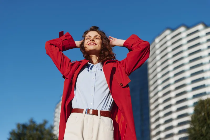Young woman with curly hair wears a bright red coat over a light blue shirt and white trousers, standing outdoors under a clear blue sky with tall buildings behind.