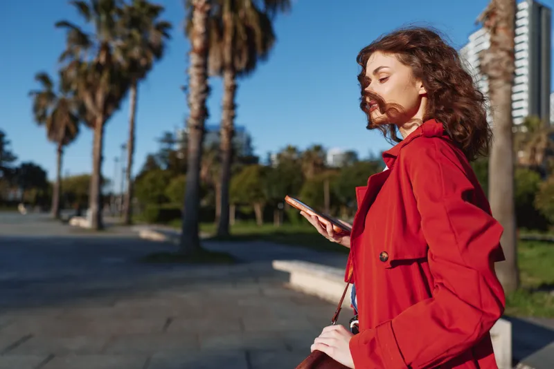 Young woman in a red coat walks outdoors on a sunny day while looking at her smartphone with a calm and focused expression.