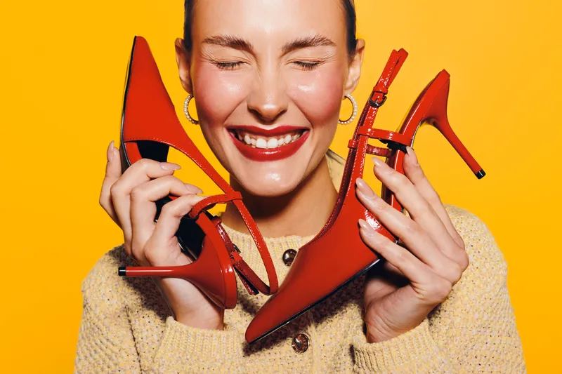 Happy woman with red lipstick holds red high heels near face, eyes closed, smiling. Bright yellow background and cozy beige sweater create warm, joyful mood of fashion and style.