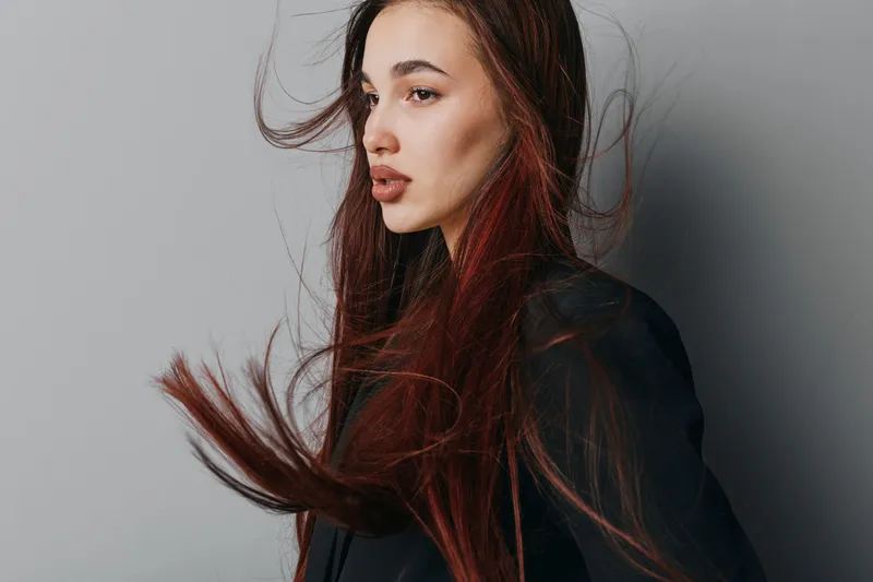 Woman with long brown hair wearing black jacket looking sideways with hair flowing against gray background in studio. Portrait of young female model with natural makeup and serious expression.