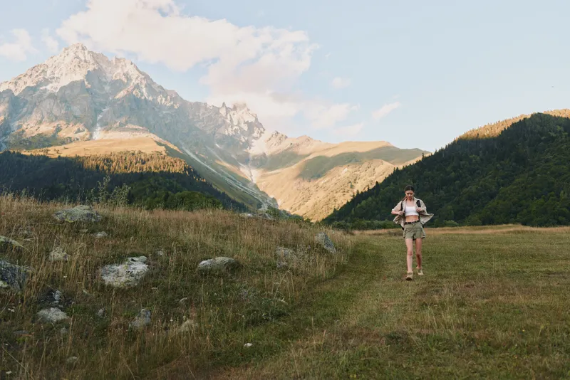 Hiker woman in a meadow with mountains in the background, carrying a backpack, exploring outdoors. Natural scenery and wellness themes with a bright, adventurous mood.