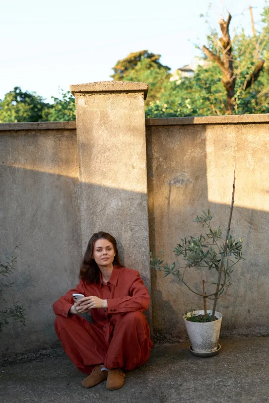 Woman in red sits on the ground beside a weathered wall, holding an object. Casual outdoor lifestyle shot emphasizing calm, simple selfcare and quiet focus