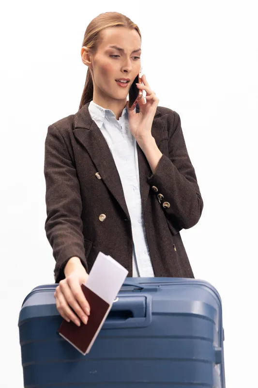 Businesswoman with suitcase and passport talks on phone, showing stress or urgency. Travel concept with luggage, documents, and communication on white background.