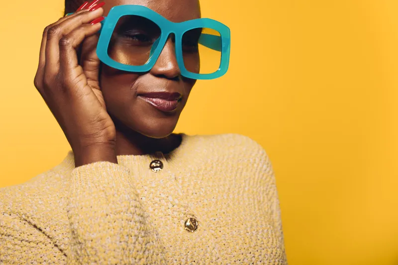 Portrait of a confident black woman with dark skin wearing large blue glasses and a beige textured sweater on yellow background. Concept of fashion, style, and modern beauty.