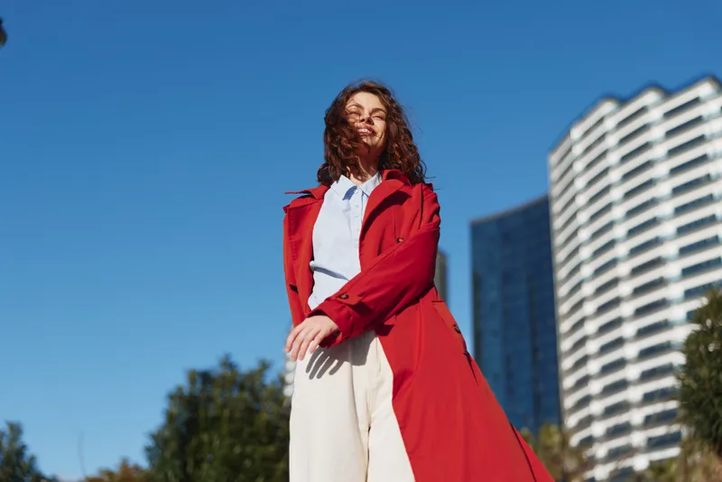 Young woman with curly hair wears a red trench coat and white pants while standing outdoors against a blue sky and modern buildings, feeling confident and relaxed.