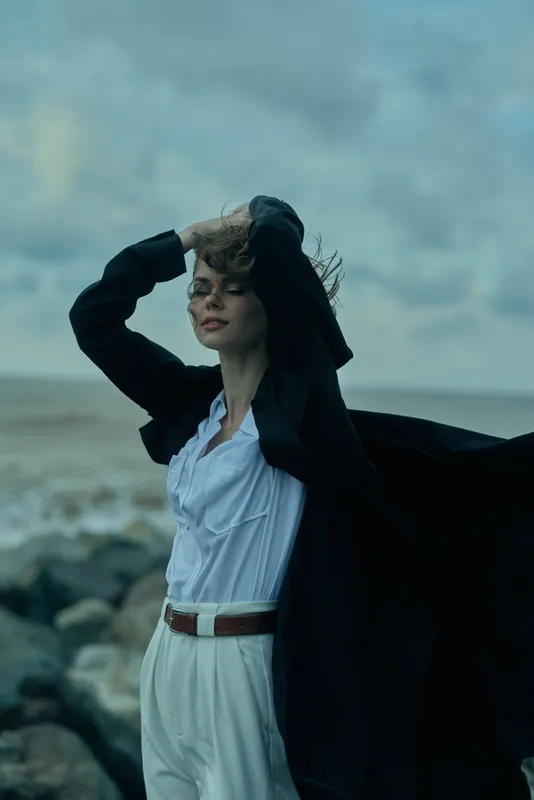 Fashion model on a windy seashore wearing a white blouse and flowing cape, belt and pants peek through, moody atmospheric scene with coastal breeze and dramatic light