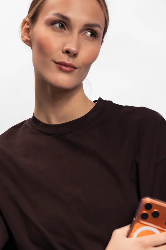 Young woman in a dark shirt holding a smartphone looks aside with a thoughtful expression. Clean background and casual style create a modern lifestyle concept.