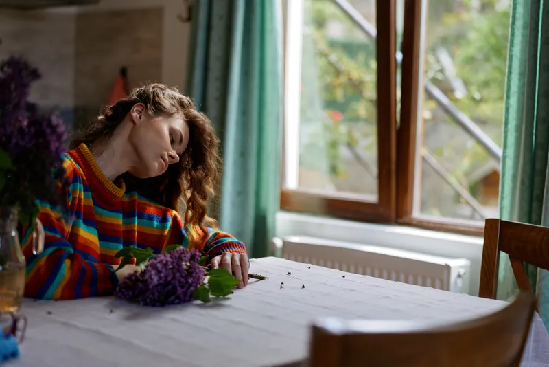 Relaxed woman with a colorful striped sweater resting at a table with purple flowers, soft natural light from a window, creating a cozy home mood and quiet self care feel.