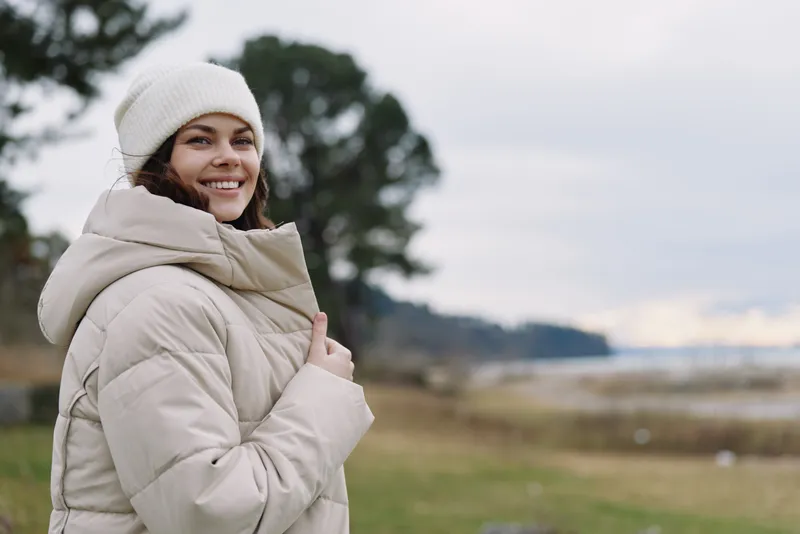 Woman in a warm winter coat and beanie smiles outdoors in a windy park, showcasing cozy attire, natural mood and seasonal comfort in a spacious landscape
