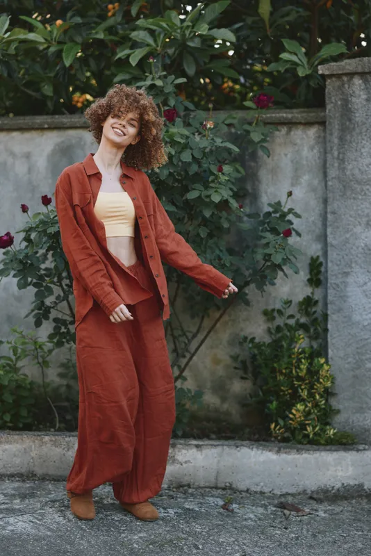 Smiling woman with curly hair wearing a rust-colored suit and pale yellow top, standing outdoors beside a stone wall. Fashion lifestyle, casual confidence, warm mood