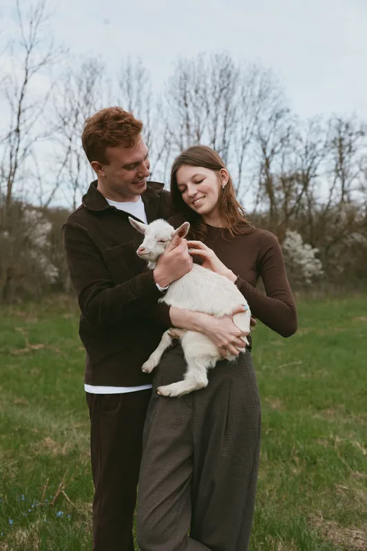 Two smiling adults cradle a young goat kid in a grassy field, showcasing farm lifestyle, tenderness, and connection with animals in natural outdoor light, creating a warm, wholesome mood and relaxed