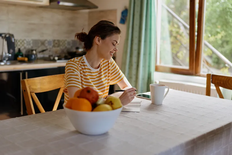 A young woman sits at a bright kitchen table writing in a notebook, smiling softly as a bowl of fruit sits nearby and warm daylight fills the cozy room.