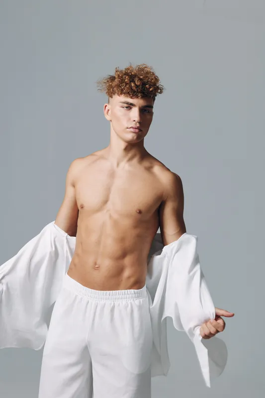 Young man with curly hair posing shirtless while removing white jacket in studio with neutral background showcasing toned body and confident expression casual fashion model.