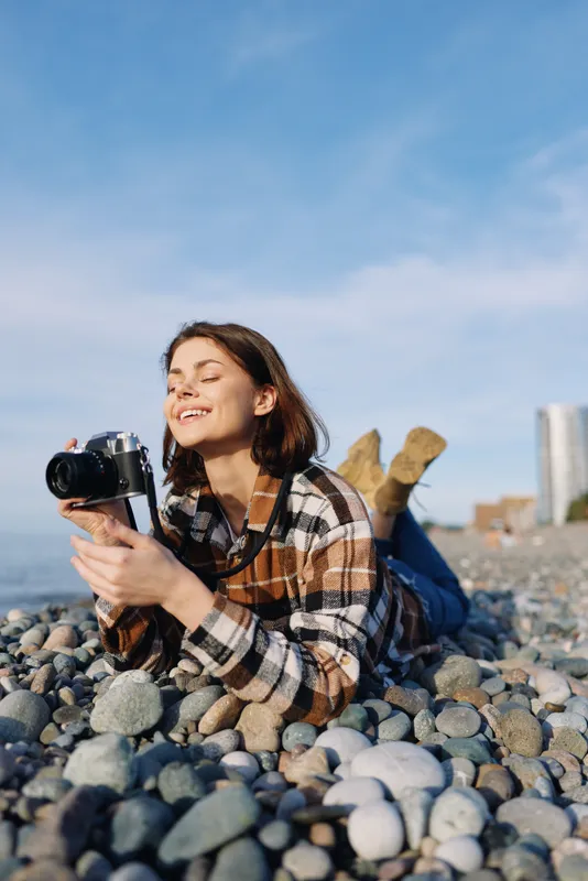 Woman photographer lying on a pebble beach, smiling and holding a camera, plaid shirt, blue sky, casual outdoor shoot capturing candid moments and natural seaside vibe