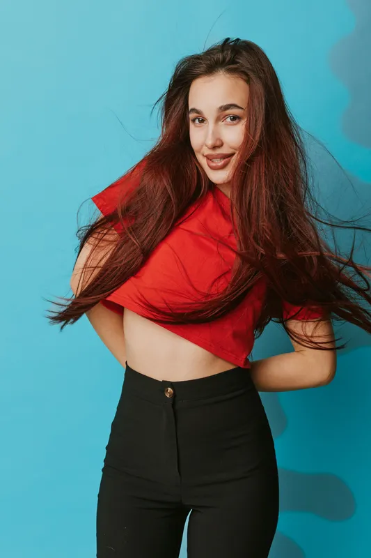 Young woman with long hair wearing red shirt and black pants posing with hands behind back on blue background. Casual style, smiling face and confident posture in studio lighting.