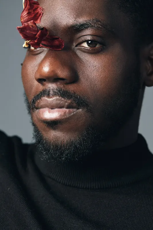 African man with dark skin and beard in creative conceptual style closeup portrait with dried red flower on face wearing black turtleneck against gray background.