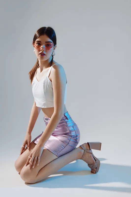 Young woman in a white crop top and shiny skirt kneels on the floor wearing sunglasses and heels. Studio shot with soft lighting. Concept of woman beauty fashion and style.
