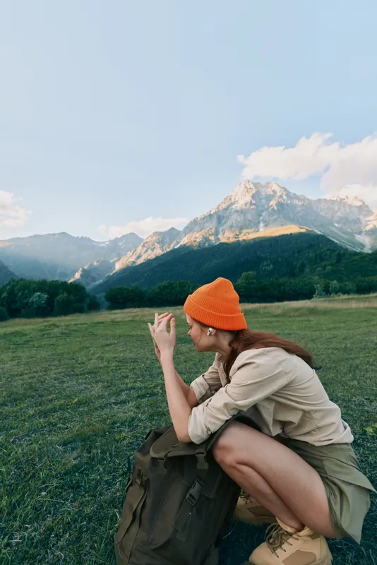 Woman with backpack sits in a meadow beside mountains, wearing an orange beanie. Closeup describes nature, hiking, and travel vibes with a calm, sunny mood.