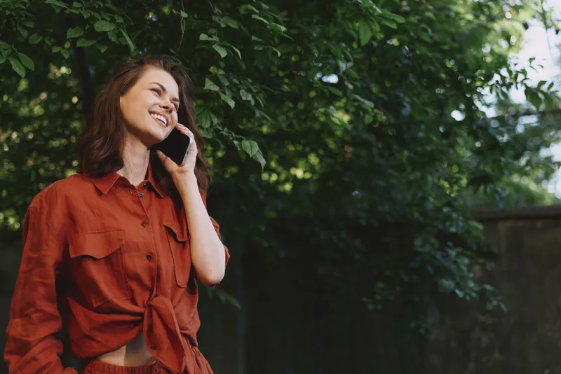 Woman lifestyle outdoors, smiling while talking on a smartphone, casual red shirt, sunlit garden setting, relaxed posture, natural colors, greenery backdrop, candid everyday moment