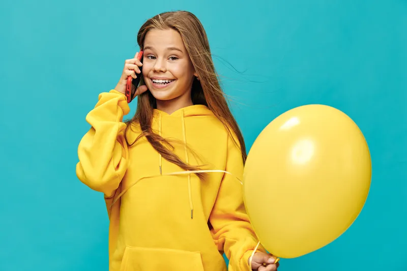 Girl in yellow hoodie talking on smartphone and holding yellow balloon on blue background smiling happy child with long hair posing isolated in studio portrait.