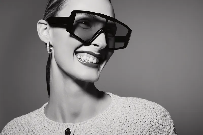 Smiling white woman with pale skin wearing oversized sunglasses and knitted sweater looks sideways. Black and white studio portrait with fashionable accessories and joyful mood.