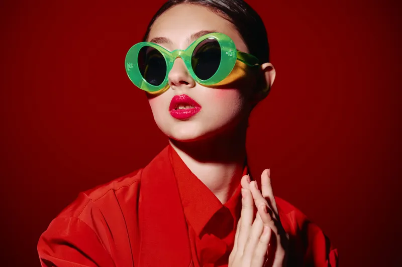 A stylish young woman wears oversized green sunglasses and vibrant red lipstick with a matching outfit. Fashion creative beauty portrait on a bold red background.