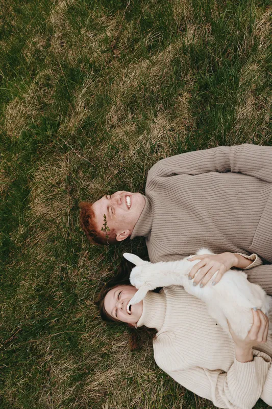 Lifestyle couple lying on grass in cozy sweaters, cuddling a white goat kid under soft light, outdoors scene showing serenity, warmth, affection and a natural wellness mood for a calm lifestyle