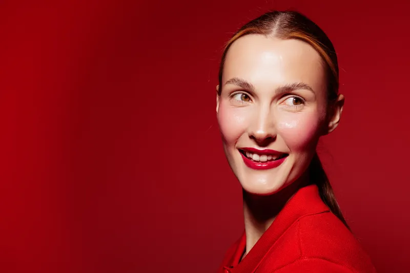 Smiling woman with red lipstick and red sweater looks sideways on red background. Portrait of happy female model with natural skin and sleek hairstyle. Concept of beauty and fashion.